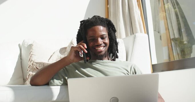 Phone ringing, African American man holding phone, smiling and checking laptop at home for work