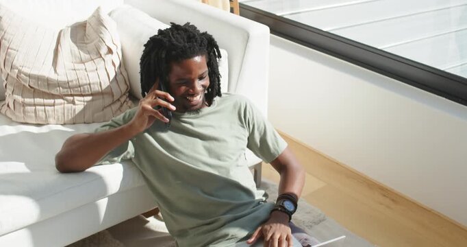 African American man answering incoming friendly call, typing on laptop and smiling on rug by sofa