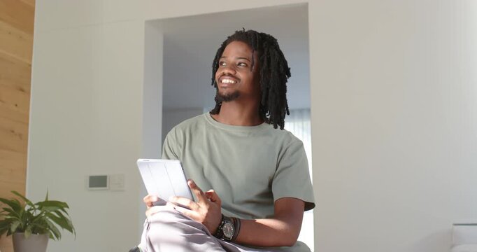 Adult African American man sitting on bed holding tablet with folio smiling, responding to right