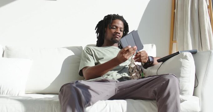 African American man sitting on sofa at home, seeing cable tangle and plugging earbuds into tablet