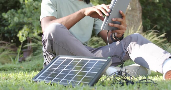 African American man sitting crosslegged under tree sun powering panel charging and tapping tablet