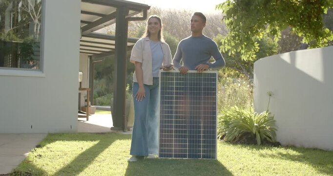 Diverse couple following woman's pointing with device, steadying solar panel on lawn, assessing sun