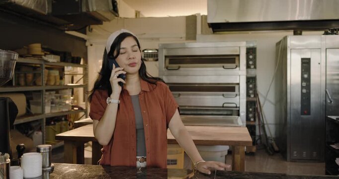 Mid adult woman taking friendly call at bakery counter, holding smartphone shifting pose, smiling