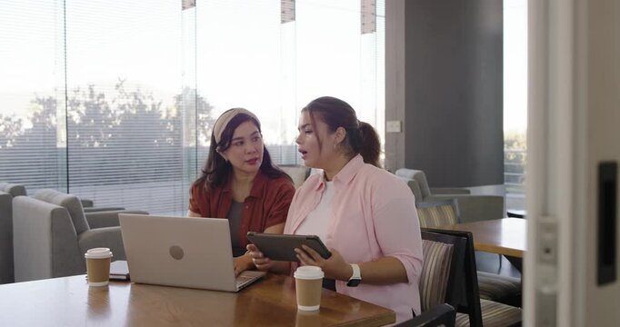 Two women reacting while camera pulling focus, tapping tablet typing laptop in lounge checking docs