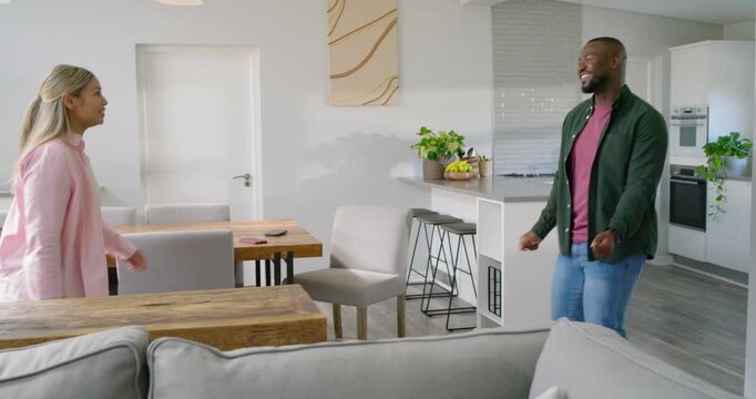 Diverse couple dancing after man gesturing, woman mirroring for bonding at home by dining table