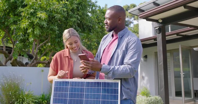 Diverse couple checking install info on phone in yard, holding solar panel and planning roof mount