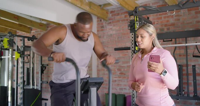 African American trainer timing on pink phone, cueing client pedaling interval on air bike in gym