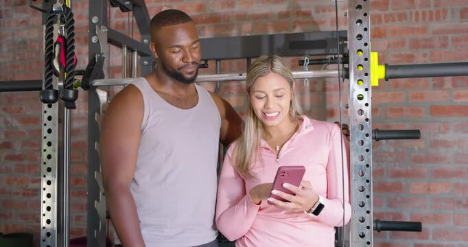 Couple at power rack, woman in pink pullover scrolling phone showing clip, man pointing, laughing