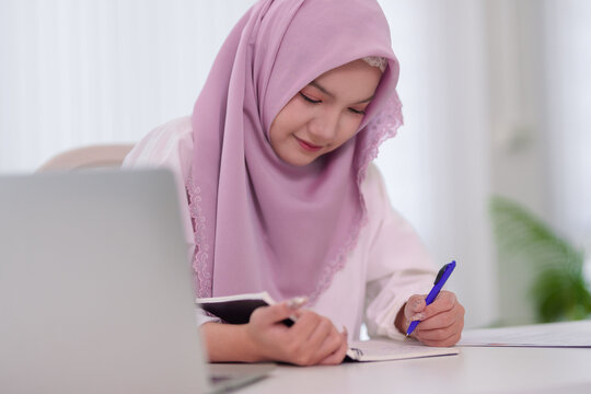 young Muslim women writing notes studying online using laptop computer smartphone for education learning business working from home office desk Islamic student