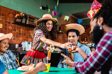 Fototapeta premium Happy Brazilian friends and family celebrating Festa Junina with traditional food and drinks, wearing straw hats and colorful costumes during the June Festival celebration.
