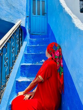 Woman in red traditional djellaba and floral headscarf sitting on blue painted stairs in Chefchaouen Morocco