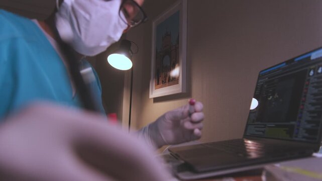 Scientist in a lab coat examining a blood test tube while working on a laptop computer