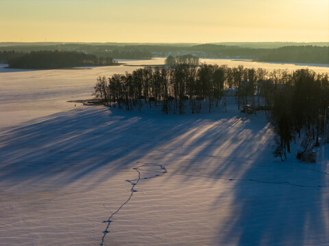 Aerial view of a frozen lake and a snow-covered island with bare trees casting long shadows during golden hour Lithuania.