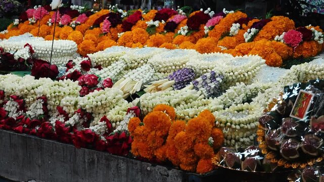 Colorful flower garlands including Jasminum sambac and Tagetes resting on wooden table, vibrant outdoor Asian market setting, cultural travel b-roll, Thai tradition background