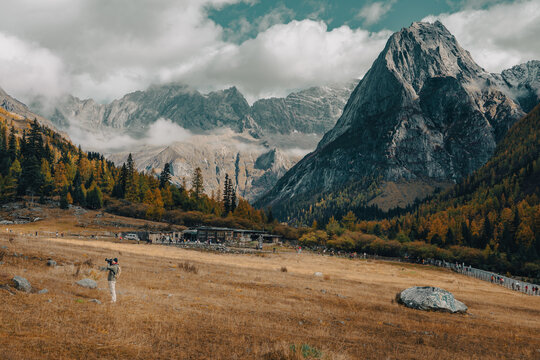 A lone photographer stands in a vast golden meadow capturing the dramatic mountain scenery at Siguniangshan (Four Sisters Mountain) National Park in Xiaojin County, Aba Tibetan and Qiang Autonomous Pr