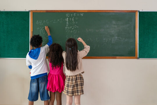 Diverse elementary students standing at classroom chalkboard solving math equations together, learning teamwork and developing educational skills in school.
