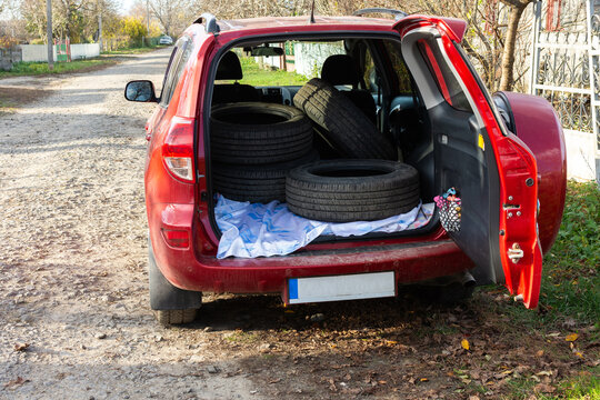 A red vehicle is parked on a gravel road while winter tires are loaded in the back. It is fall, and the trees are colorful with leaves. The vehicle shows signs of tire changing activity