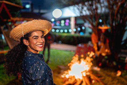 Happy young woman in traditional caipira costume with straw hat smiling at a Festa Junina celebration at night near a bonfire.
