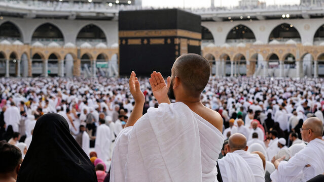 Muslim pilgrim in white ihram clothing praying with raised hands facing the Holy Kaaba during Hajj or Umrah pilgrimage at Masjid al-Haram in Mecca, Saudi Arabia.