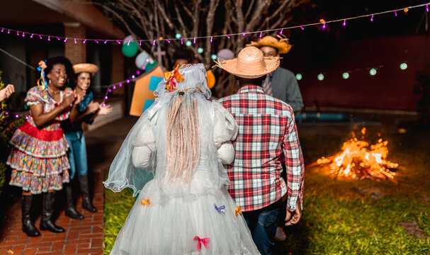 Traditional Brazilian Festa Junina celebration featuring a mock wedding with a bride and groom, bonfire, and guests in caipira costumes at night.