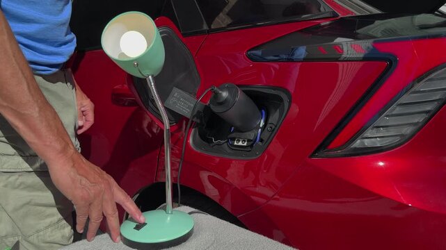 Man's hand operating a switch to illuminate a domestic lamp plugged into a vehicle to load adapter on a red EV electric car 