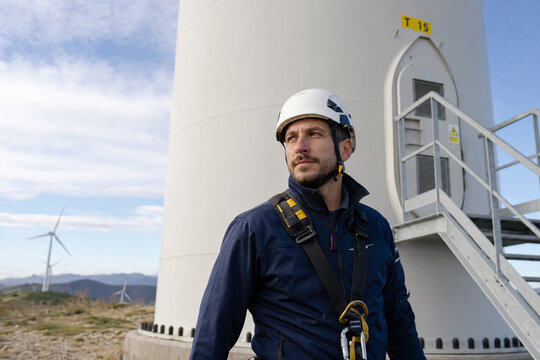 Professional male wind turbine technician wearing a safety helmet and harness stands at the base of a large wind generator on a bright day at a renewable energy power station.