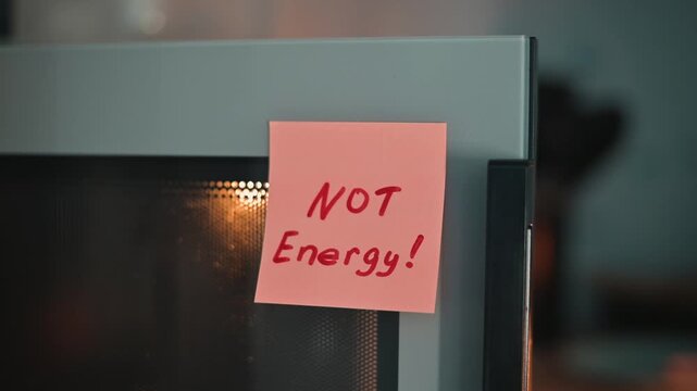 A hand reaches for the microwave door in a kitchen setting, where sticky notes serve as reminders about energy use. The scene highlights the importance of mindful cooking practices during busy times