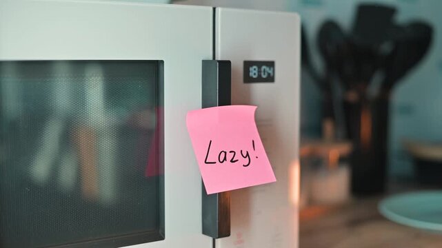 A hand places a pink sticky note LAZY on the door of a microwave. This action highlights the importance of reminders in everyday kitchen tasks to motivate individuals