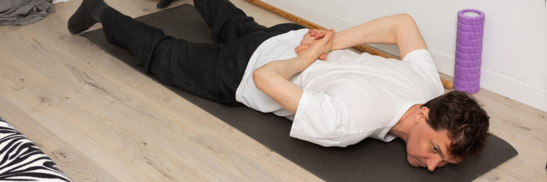 A man, 53 years old, lies on a yoga mat at home, performing a stretching exercise. He focuses on his fitness routine in a simple indoor space with plants around, banner