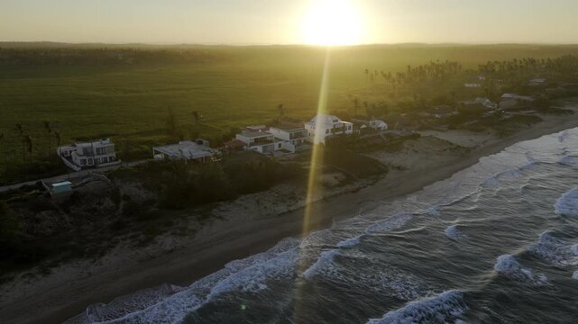 Drone facing sunset, flies backwards at angle along beach and over trees and house in Tofo, Mozambique