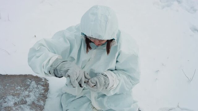 Survivor repairing sensor in snow, focused hands wiring small device while hooded parka and mittens show frost and wear improvised repair, salvage mentality, abandoned station edge, tense quiet