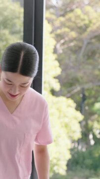 Vertical video: Smiling caregiver in pink scrubs guiding senior entering glass door, copy space