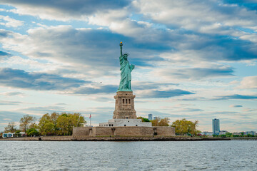 Fototapeta premium Statue Of Liberty On Liberty Island Waterfront. Statue of Liberty on Liberty Island viewed from water with cloudy sky and New York Harbor.