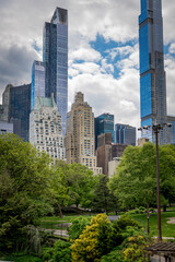 Fototapeta premium Central Park Trees Below Midtown Manhattan Towers. Central Park greenery with footpaths, lamps, and Midtown Manhattan skyscrapers under cloudy sky.