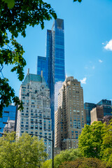 Fototapeta premium Midtown Manhattan Towers Rising Above Central Park Trees. Midtown Manhattan skyscrapers and historic buildings above Central Park trees under blue sky.
