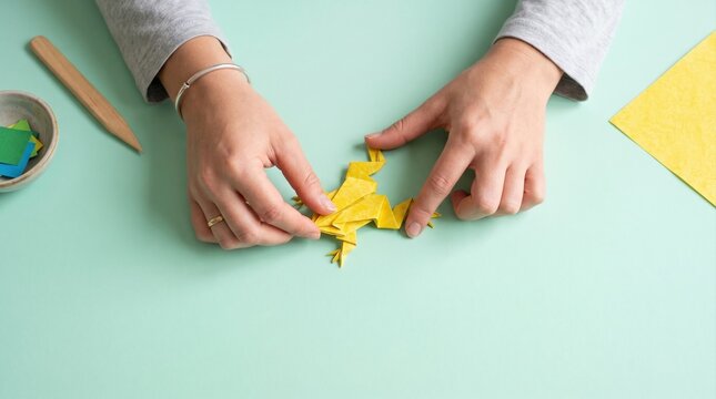 Overhead view of hands crafting a yellow origami frog from colored paper on a light green surface, surrounded by crafting tools.
