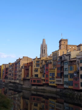 Colorful hanging houses reflected in the Onyar River with the historic Girona Cathedral spire in the background under a clear blue sky, capturing the iconic Mediterranean urban landscape in Spain.