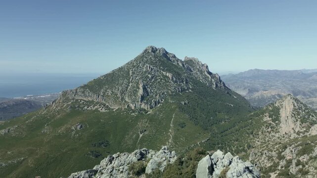 Majestic aerial view moving backward over a rocky ridge, revealing the impressive Puig Campana  (1.406 m) mountain peak against a clear blue sky, with the mediterranean sea visible in the distance.
