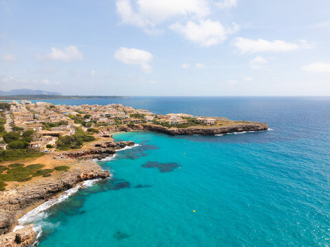 Aerial view of the turquoise coastline and rocky cliffs with residential buildings and lush vegetation under a bright sky in Sa Coma, Balearic Islands, Spain.