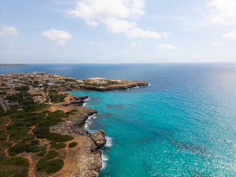 Aerial view of the coastal resort area with turquoise waters, rocky cliffs, and residential buildings under a blue sky in Sa Coma, Balearic Islands, Spain.