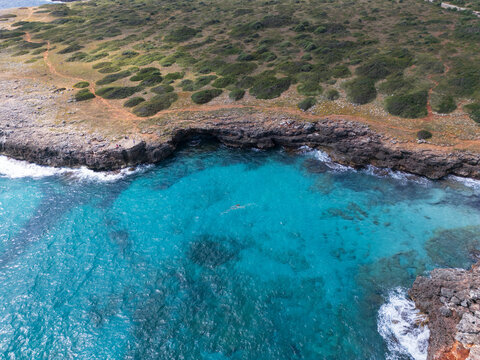 Aerial view of the rugged rocky coastline and crystal clear turquoise waters of a secluded cove with green scrubland and a dirt path Sa Coma, Balearic Islands, Spain.