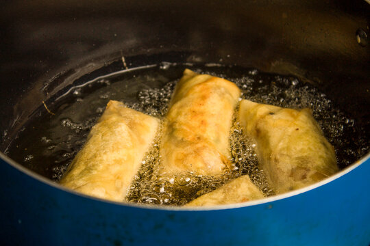 Close-up of golden brown vegetable spring rolls deep frying in hot bubbling oil inside a blue pot. 