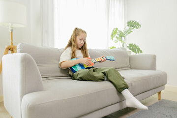 Obraz premium A young girl sits on a beige couch in a bright living room, playing a colorful ukulele with focus and calm.