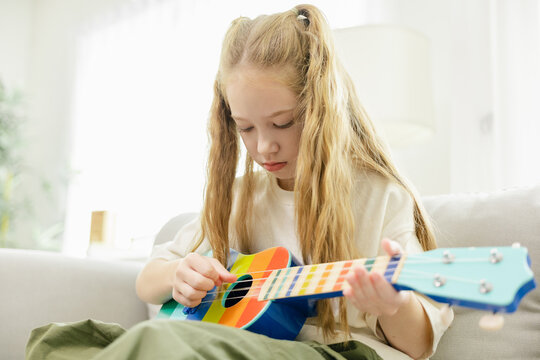 A young girl with long blonde hair plays a colorful ukulele while sitting on a sofa in a bright, cozy room.