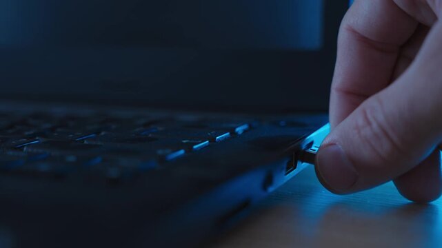 Hand inserting flash drive or thumb drive into laptop computer in dark office. Secret files being copied from server or hacked computer. Spy penetrating business or government computer with malware