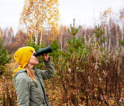 Woman looking through binocular in autumn forest. Young tourist exploring nature, watching birds and wildlife. Outdoor leisure activity, hiking travel lifestyle, exploration concept.