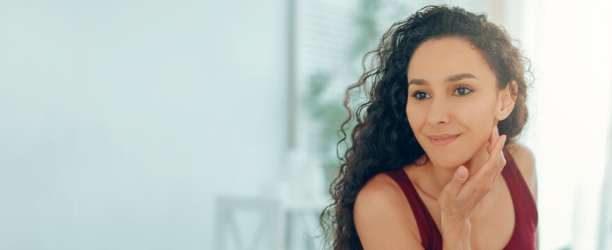 A woman with curly hair is smiling and gently touching her face. She is in a bright room with natural light and plants in the background. This scene shows a moment of self-reflection or joy.