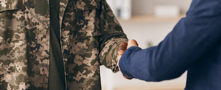 Two individuals shake hands in a room. One is wearing a military uniform while the other wears a casual outfit. They appear engaged and respectful.