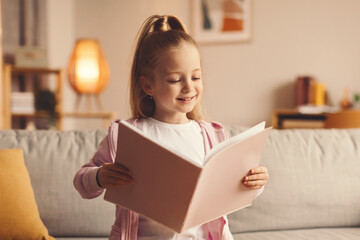 Naklejka premium A child stands in a living room holding a book with both hands. She smiles as she looks at the pages. The room has soft lighting and a comfortable sofa.