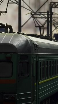 A green train travels on metal tracks in an urban location. Electric structures stand tall in the background. The sun sets, casting a warm glow on the scene.